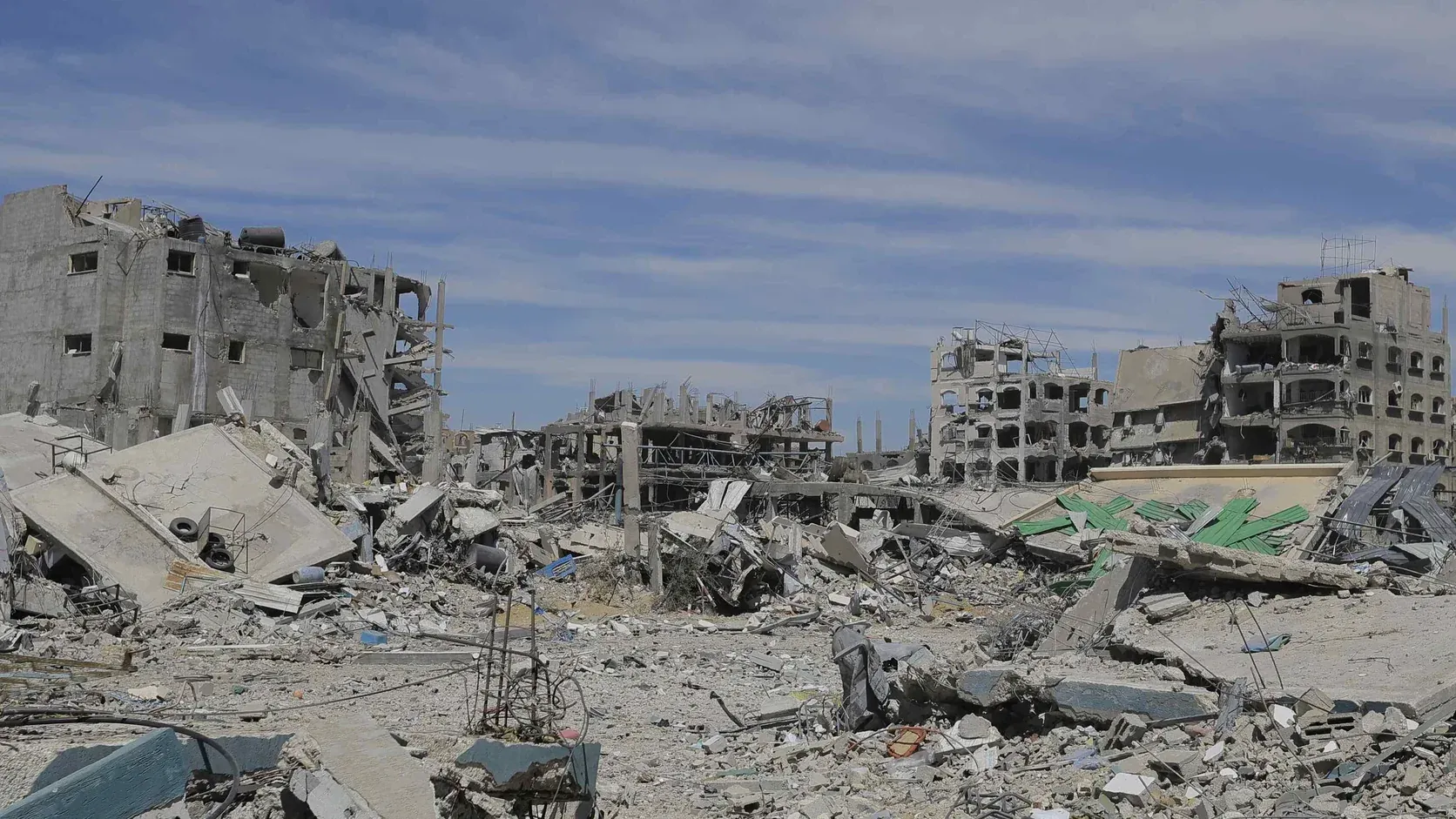 Sönderbombade flervåningshus och fält av betongbråte i flyktinglägret Jabalia i norra Gaza under en blå himmel med tunna molnslöjor.