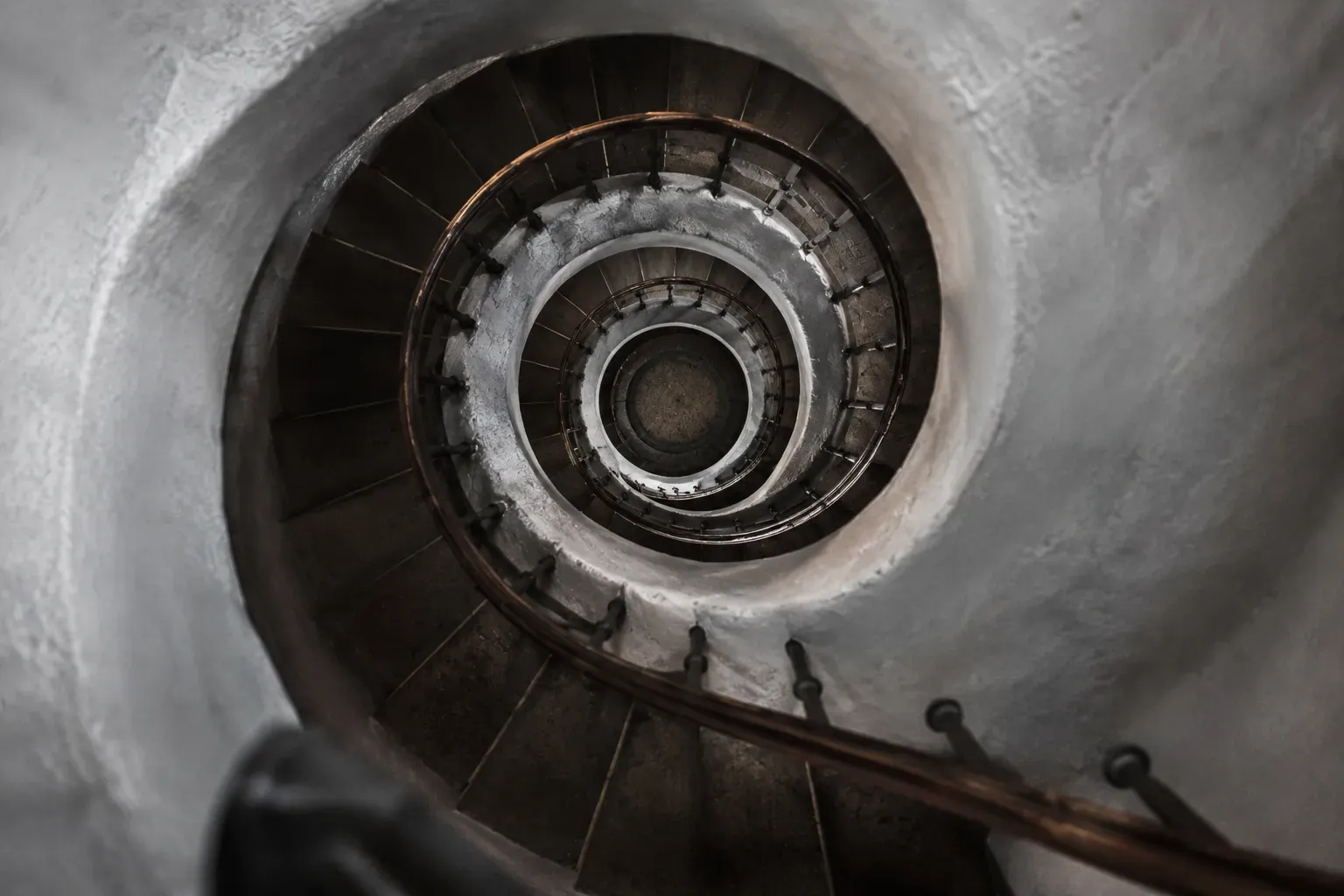 Vitkalkad spiraltrappa fotograferad uppifrån, där trappsteg och ledstång virar sig i allt tätare varv mot ett mörkt centrum.